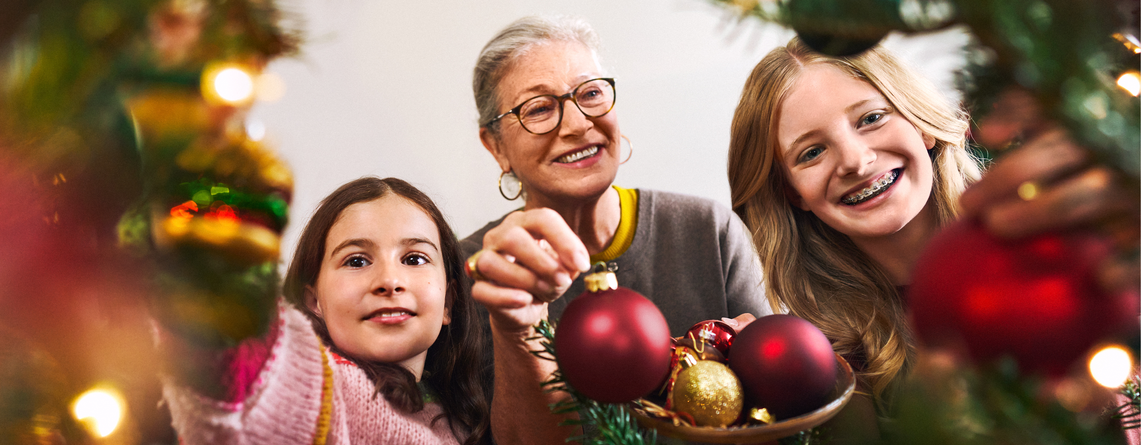 Eine Großmutter und zwei Mädchen schmücken einen Weihnachtsbaum mit roten und goldenen Kugeln.