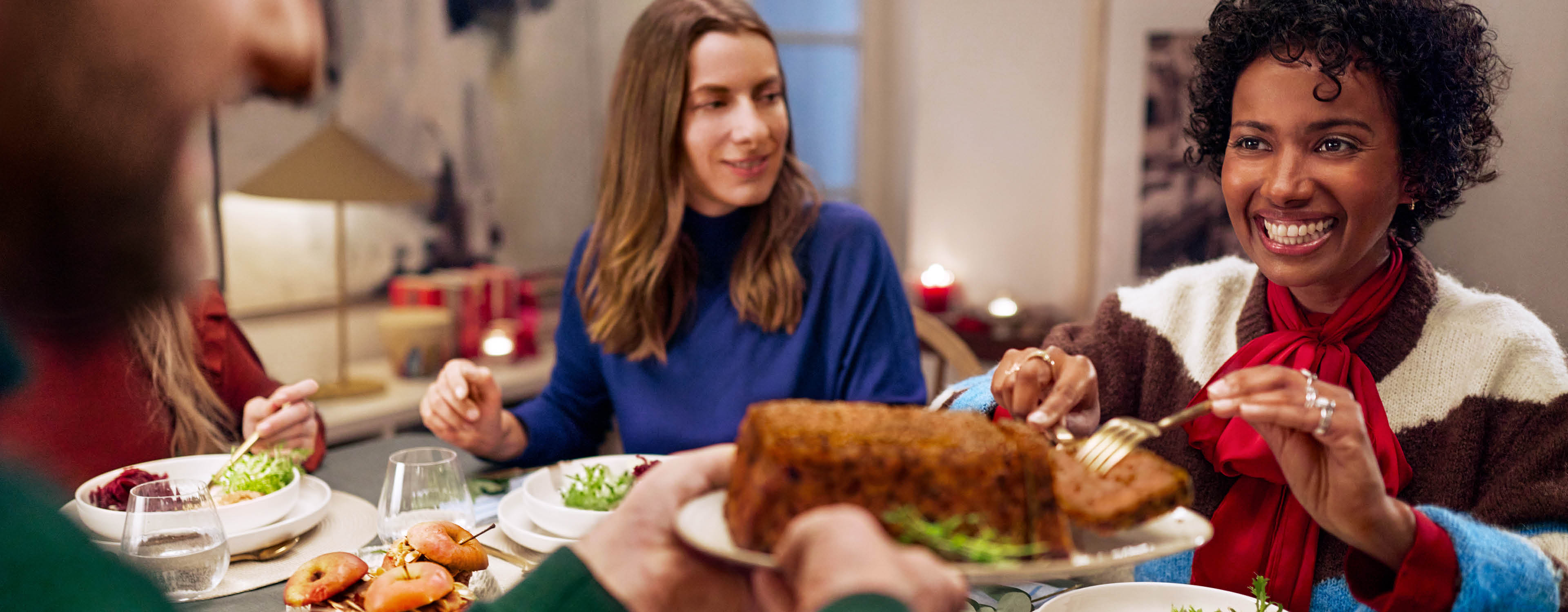 Zwei Frauen am Esstisch, eine lächelt und nimmt sich Essen von einer Platte.