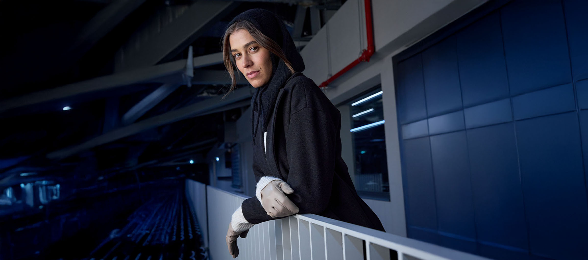 Frau in schwarzem Mantel und Handschuhen lehnt an einem Geländer in urbaner Umgebung.