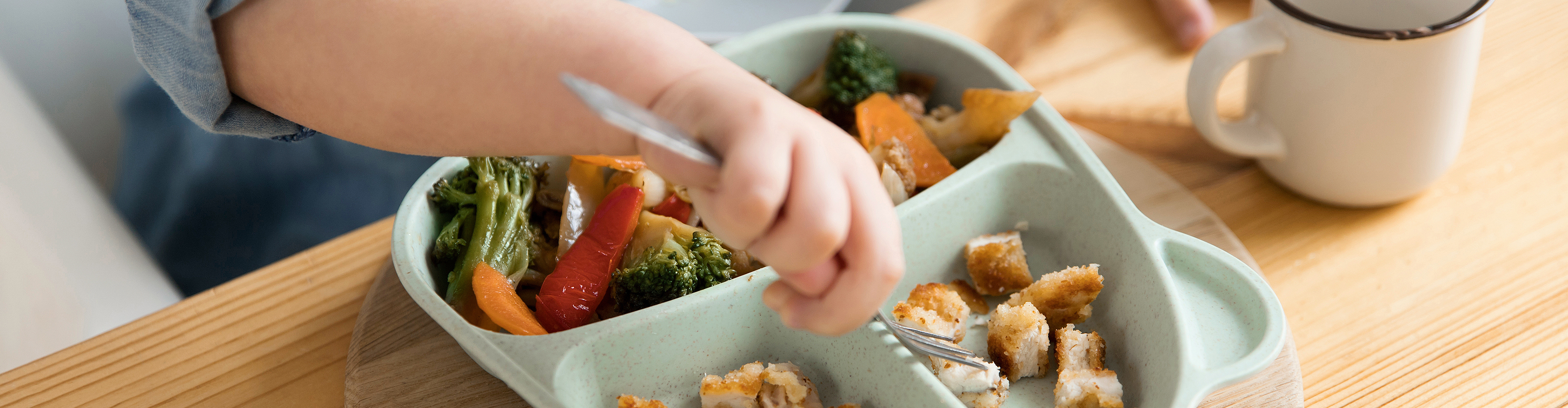 Kind isst mit Gabel aus einem Teller mit Unterteilungen, gefüllt mit Gemüse und Hähnchen.