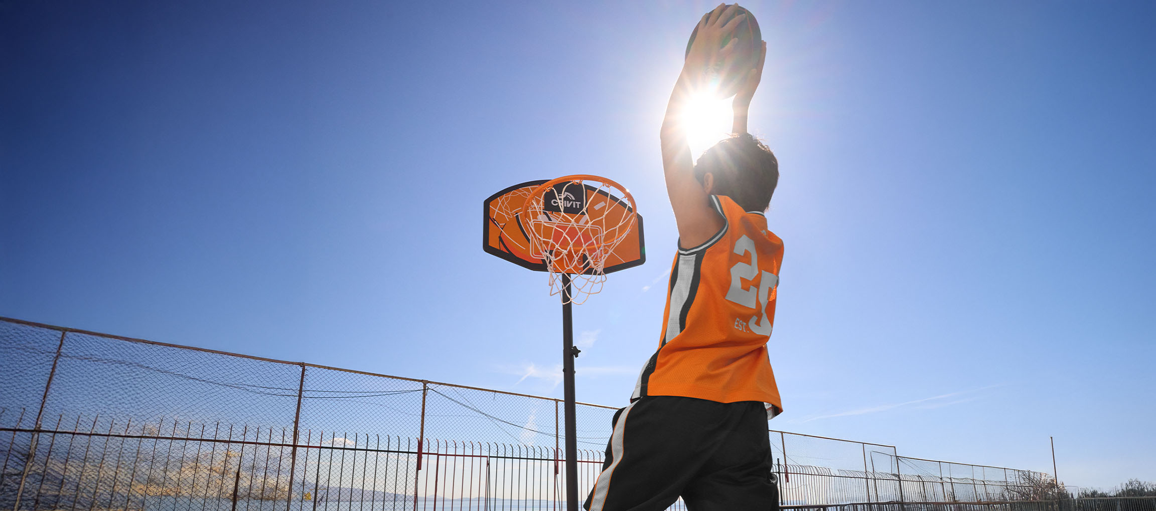 Junge im orangefarbenen Basketballtrikot wirft den Ball bei Sonnenschein in den Korb.