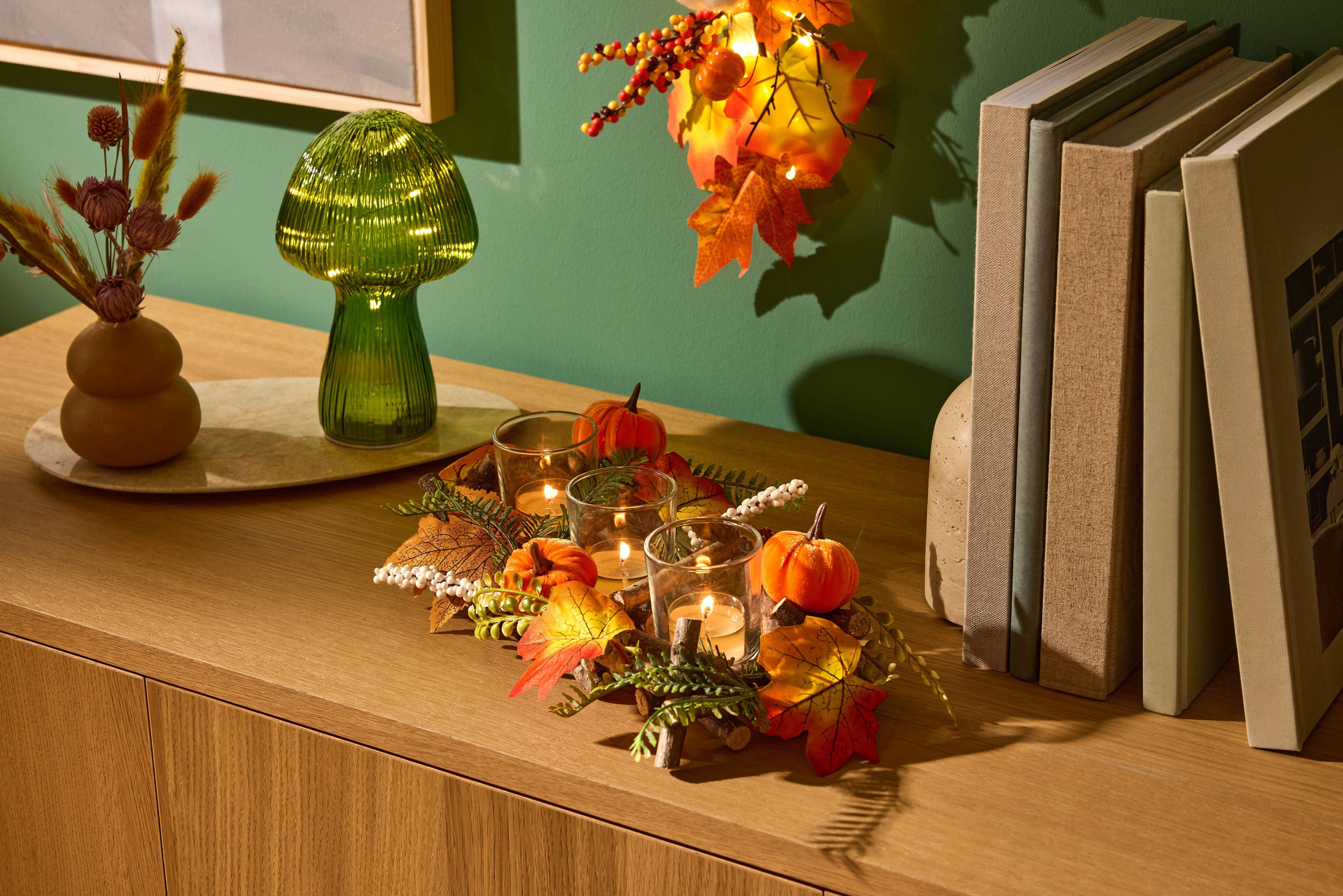 Herbstliche Tischdekoration mit Kürbissen, Kerzen und Blättern, daneben eine grüne Pilzlampe und Bücher.