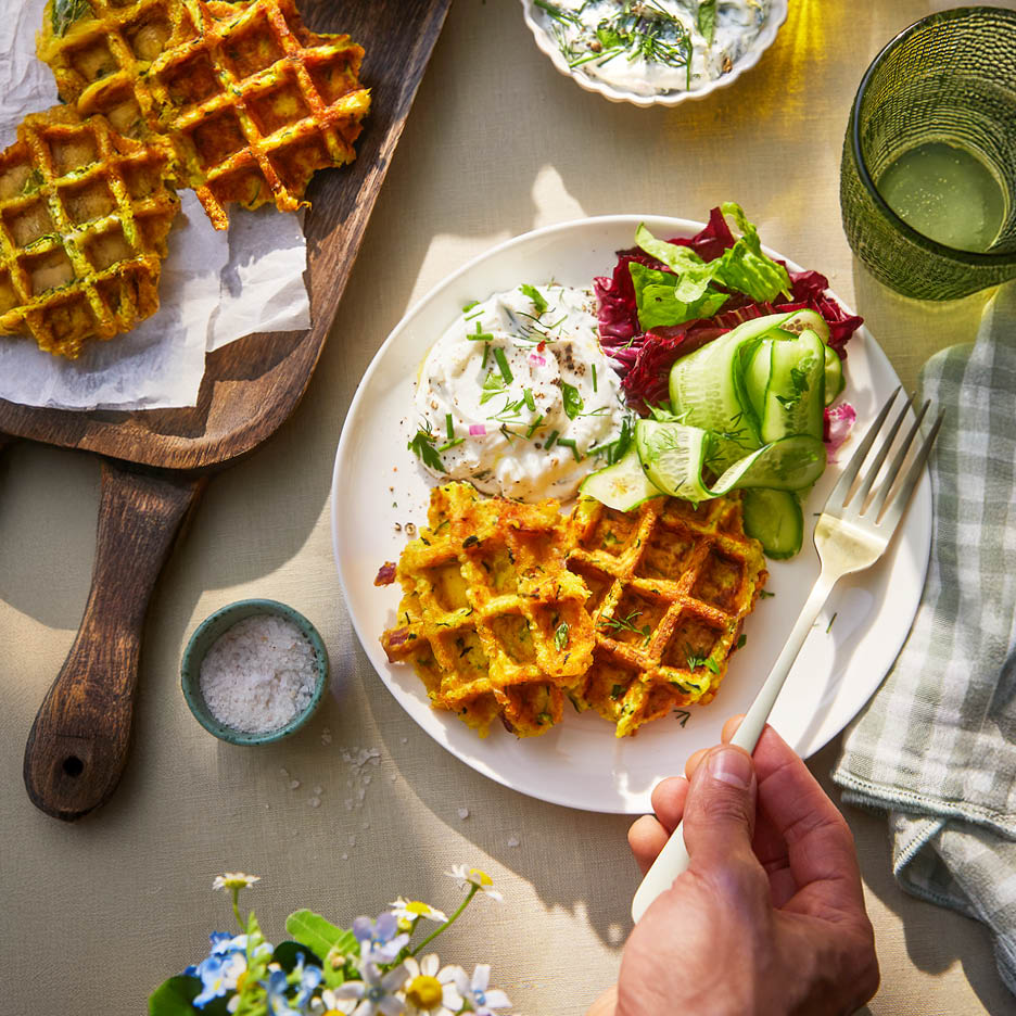 Eine Mahlzeit mit herzhaften Waffeln, Tzatziki und Salat, serviert auf einem Tisch mit Blumen.