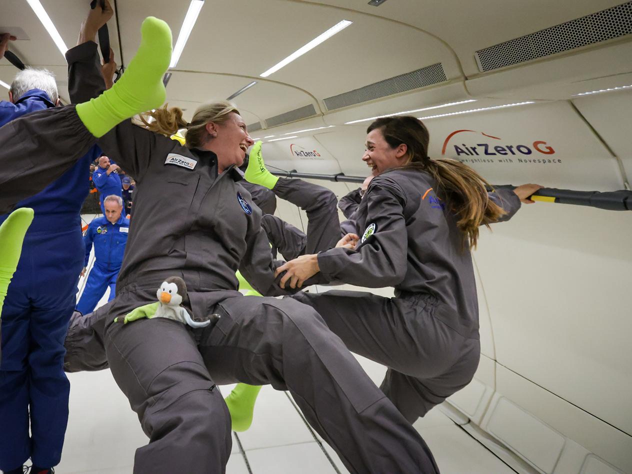 Melanie und Daniela in Overalls und leuchtend grünen Socken schweben in einem Flugzeug und lachen.