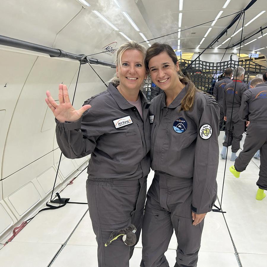 Melanie und Daniela in grauen Overalls lächeln im Flugzeug, eine macht eine Handgeste.