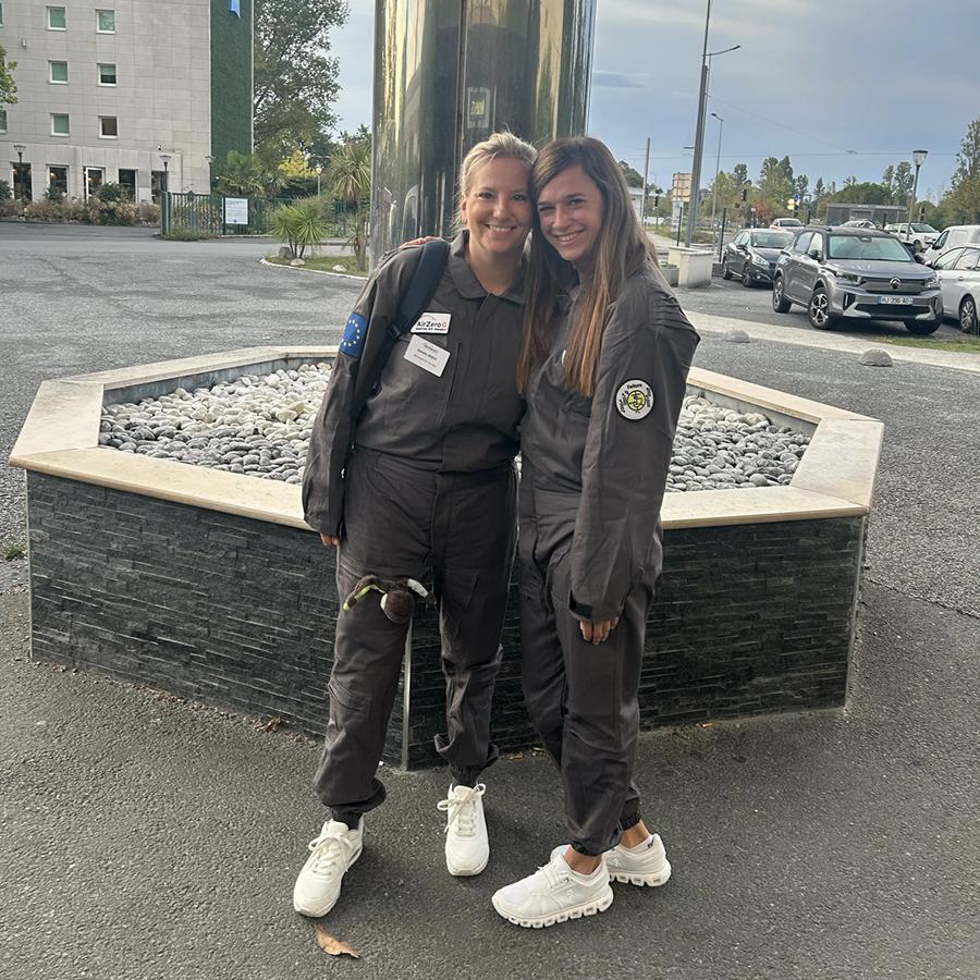 Melanie und Daniela lächeln in Overalls und weißen Turnschuhen vor einem Gebäude.