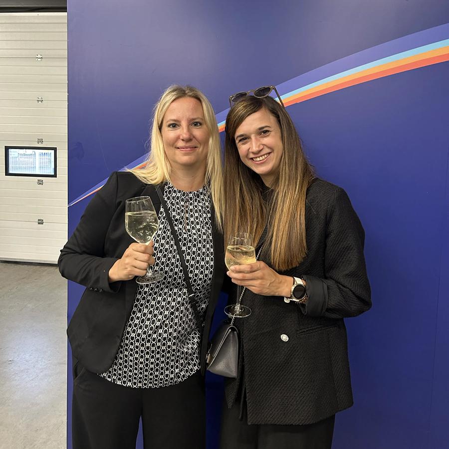 Melanie und Daniela stoßen mit Sektgläsern vor einer blauen Wand an.