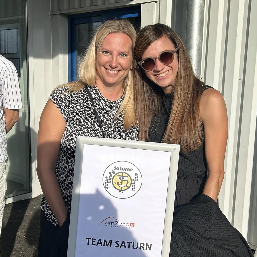 Melanie und Daniela posieren vor einem Schild mit der Aufschrift „TEAM SATURN“ und einem Logo.