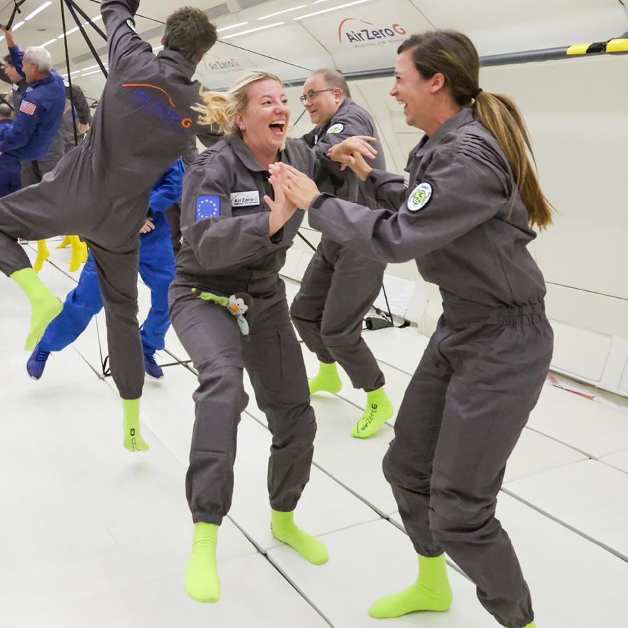 Melanie und Daniela in grauen Overalls und neongrünen Socken schweben lachend in einem Flugzeug.
