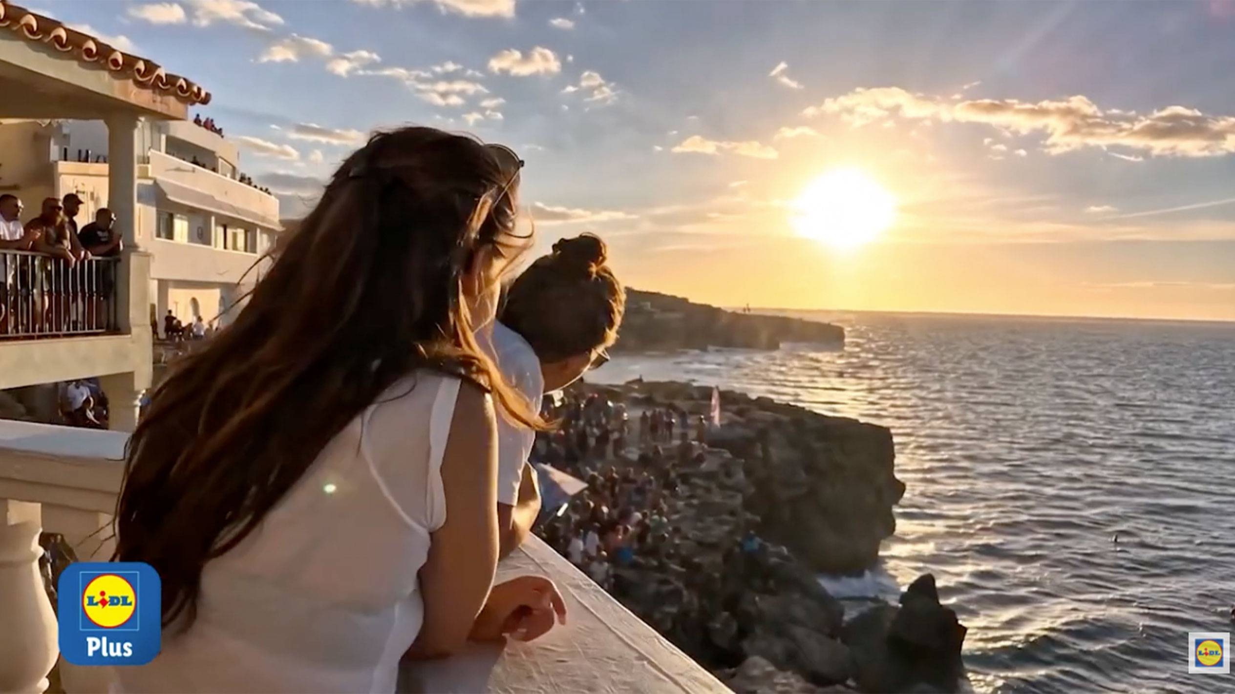 Zwei Frauen blicken von einem Balkon auf einen Sonnenuntergang über dem Meer und einer Menschenmenge.