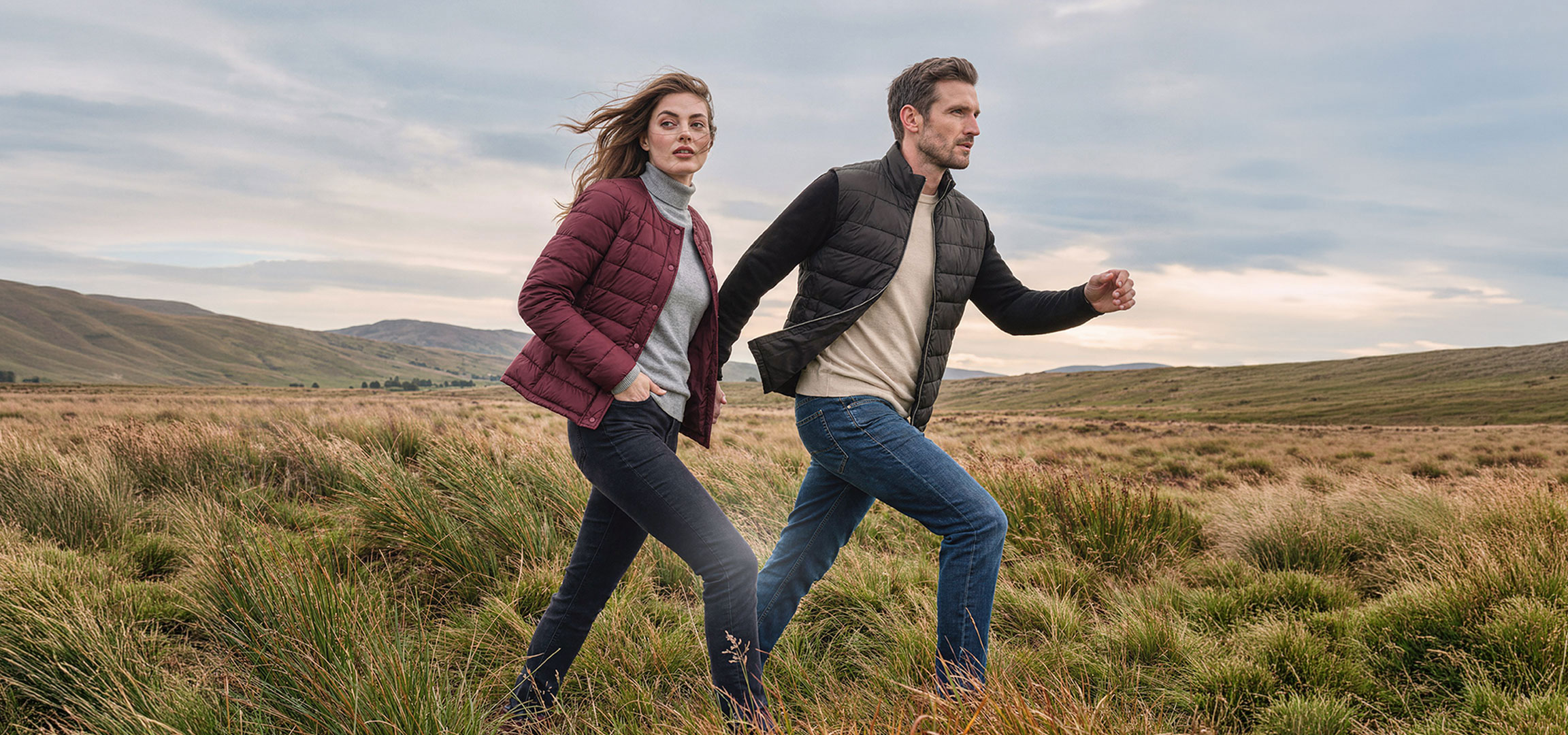KI-generiert: Paar in leichten Jacken und Jeans geht durch eine grasbewachsene Hügellandschaft.
