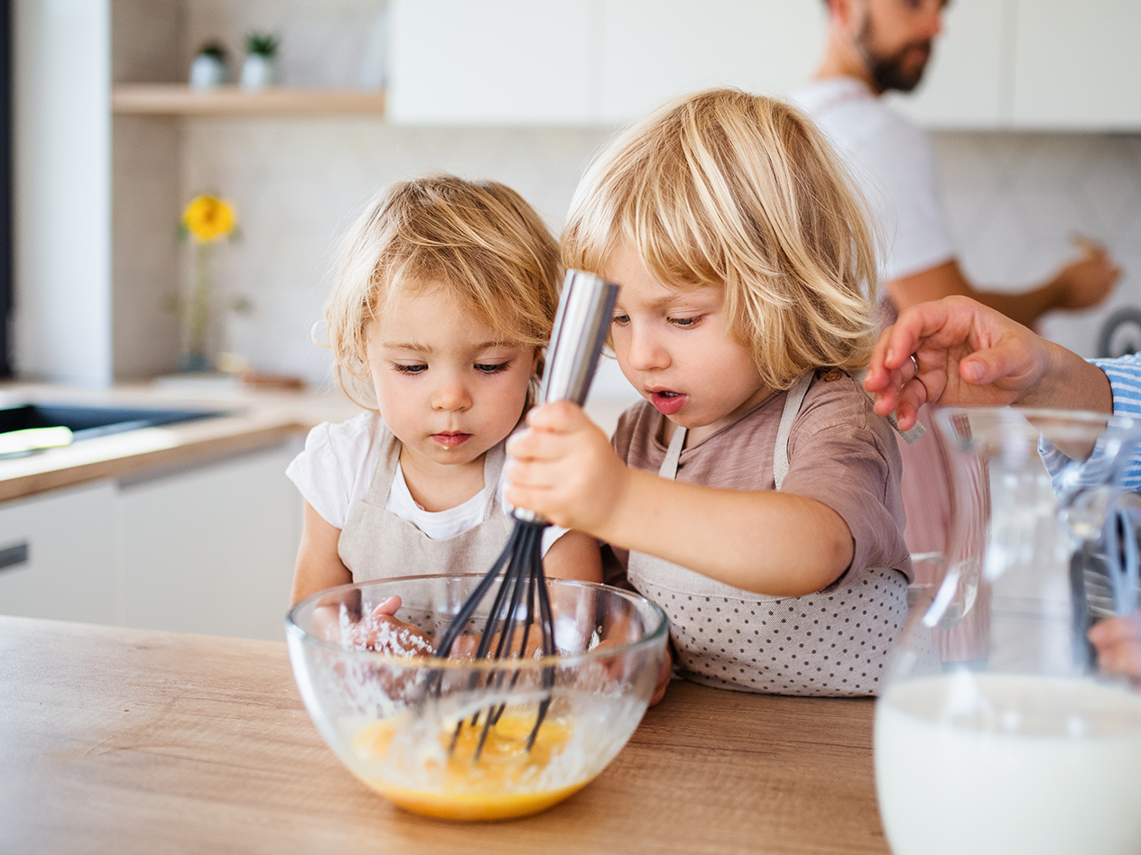 Zwei Kinder rühren in einer Schüssel mit Eiern, ein Mann steht im Hintergrund in der Küche.