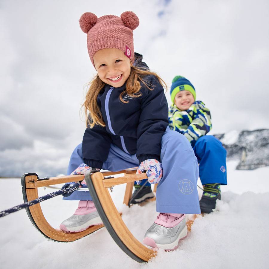 Kinder in Winterkleidung und Bommelmützen, die auf einem Schlitten im Schnee fahren.