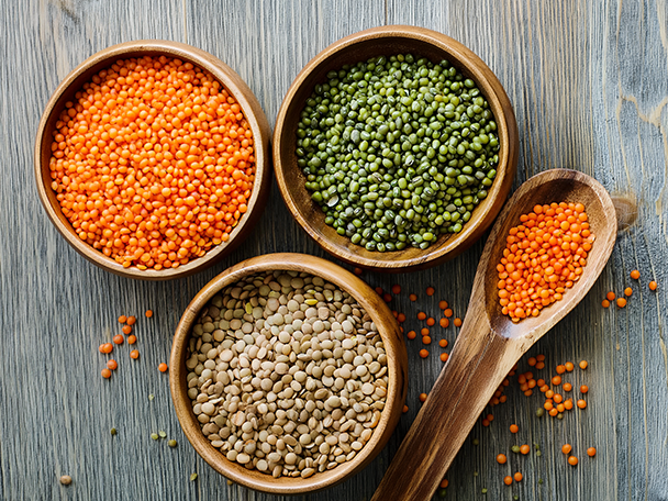Drei Schalen mit verschiedenen Linsen (rote, grüne, braune) und ein Holzlöffel auf Holz.