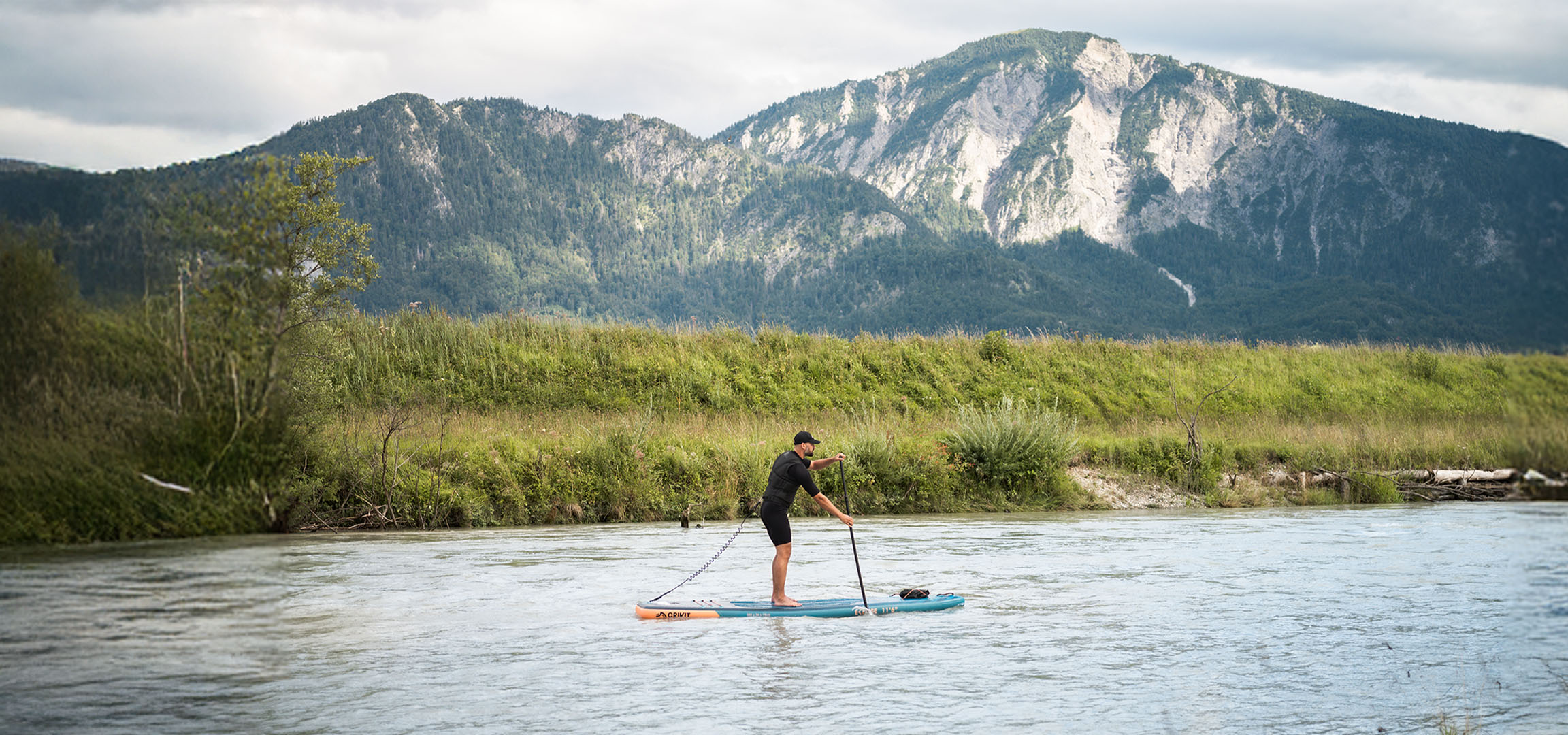 Mann auf einem Paddleboard auf einem Fluss mit Bergen und üppiger Vegetation im Hintergrund.