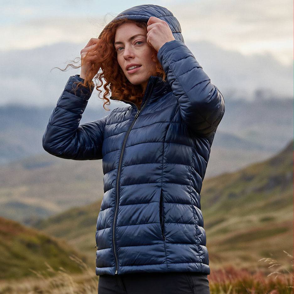 Frau mit roten Haaren in einer dunkelblauen Kapuzen-Steppjacke in einer Berglandschaft.
