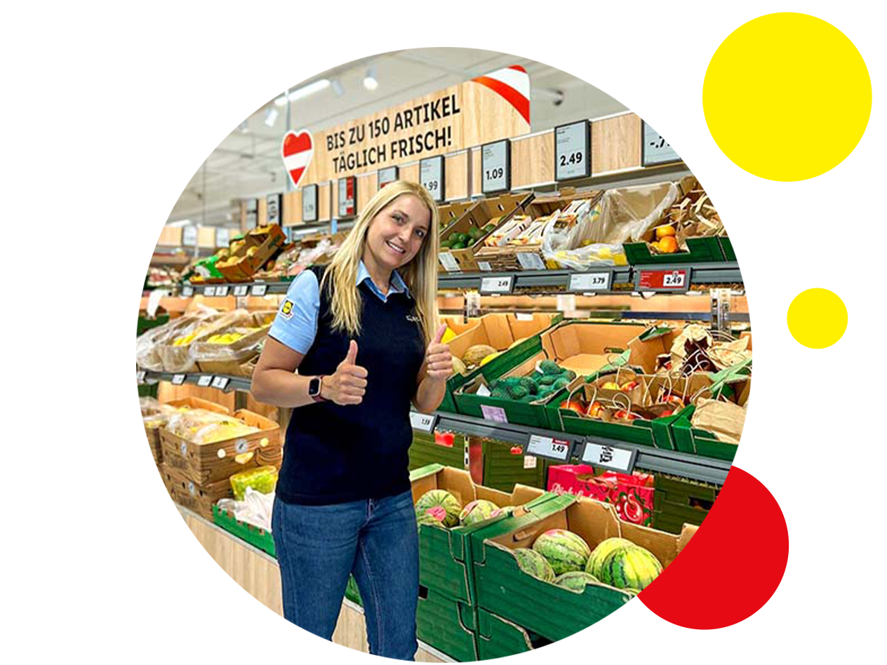 Eine lächelnde Mitarbeiterin steht in einem Supermarkt vor Obst- und Gemüseständen.