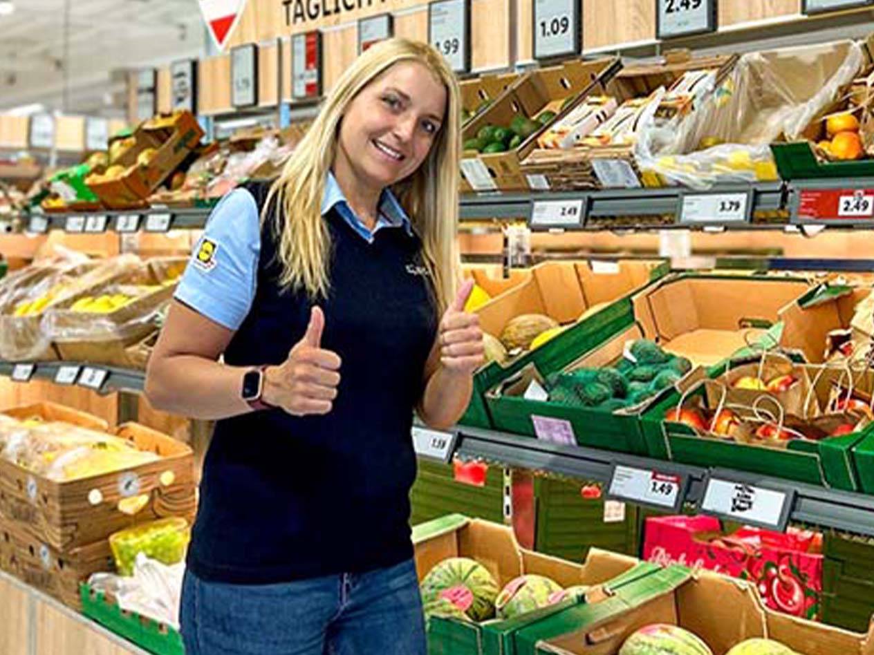 Eine lächelnde Frau gibt in einem Supermarkt mit Obst- und Gemüseständen zwei Daumen hoch.