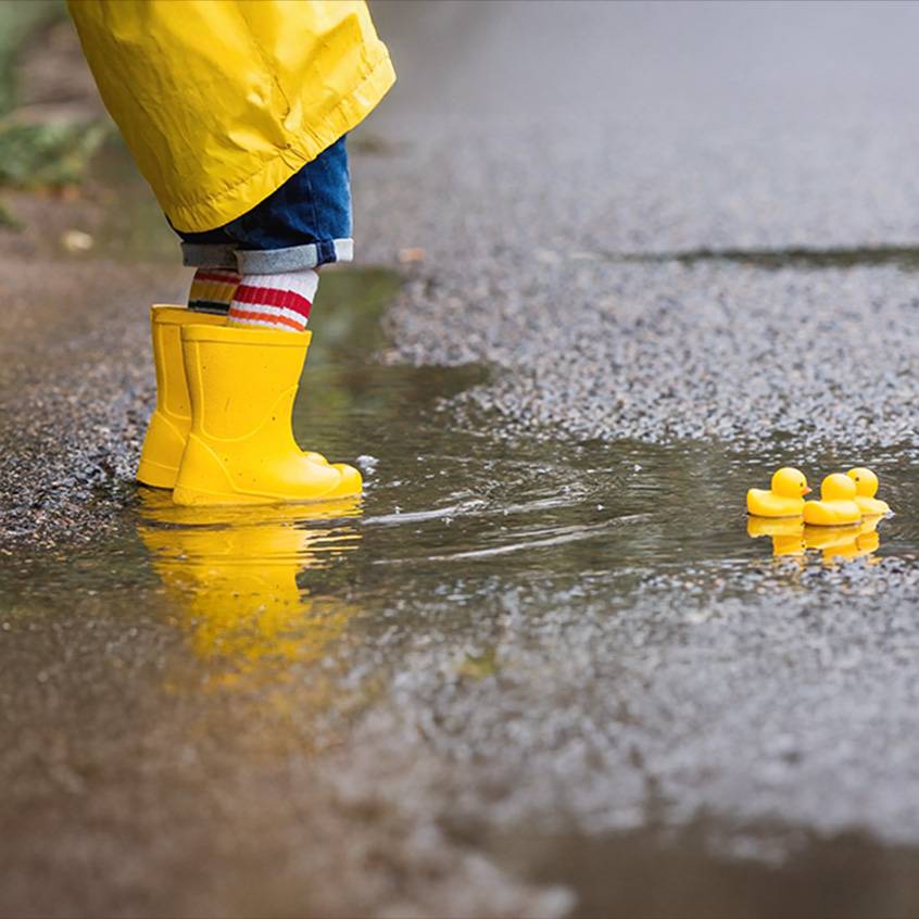 Kind in gelben Gummistiefeln und Regenjacke in einer Pfütze mit Quietscheentchen.