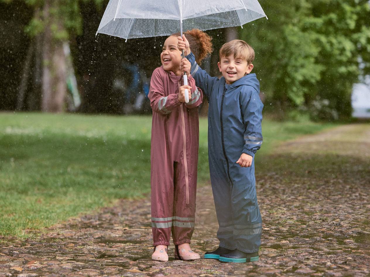 Zwei glückliche Kinder in Regenanzügen unter einem transparenten Regenschirm.