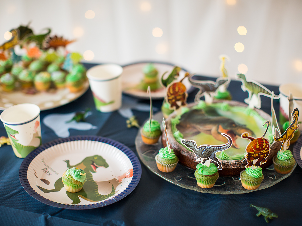 Dinosaurier-Mottoparty mit Kuchen, Cupcakes und Papptellern auf einem dunklen Tisch.