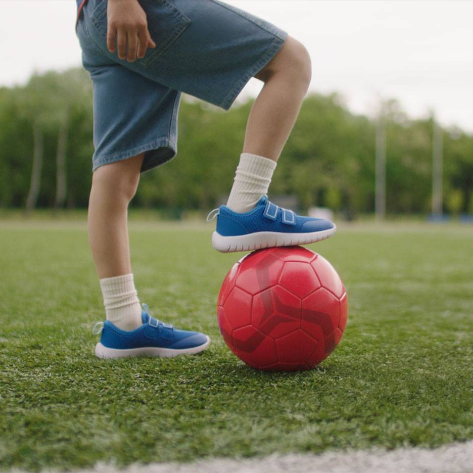 Kind in Shorts und blauen Turnschuhen, mit einem Fuß auf einem roten Fußball.