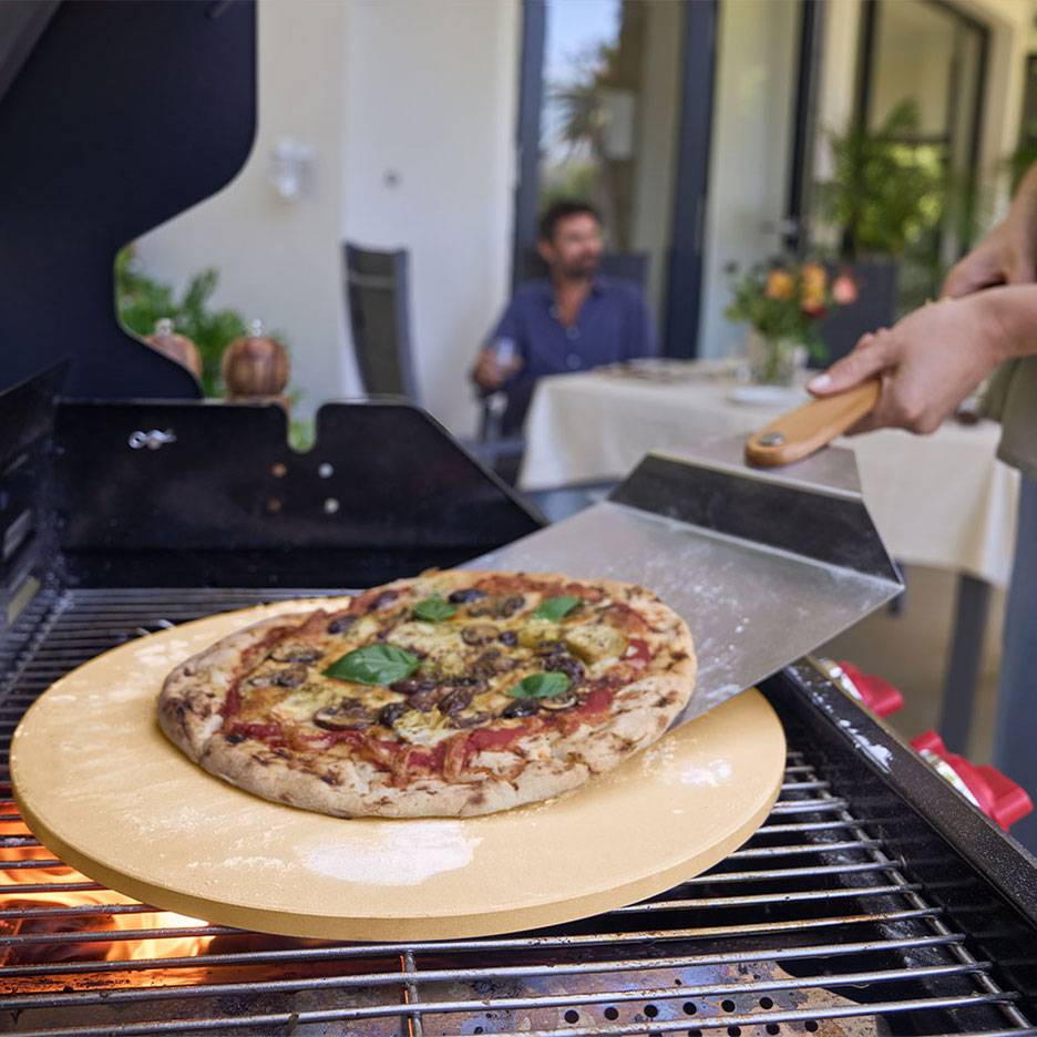Pizza auf einem Pizzastein auf dem Grill, mit einem Pizzaschieber und einem Mann im Hintergrund.