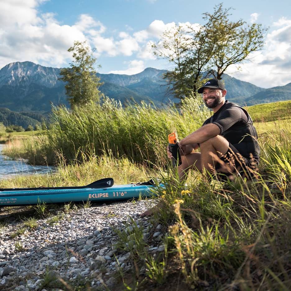 Lächelnder Mann mit Kappe und Schwimmweste, sitzt neben einem blauen SUP-Board am Fluss.