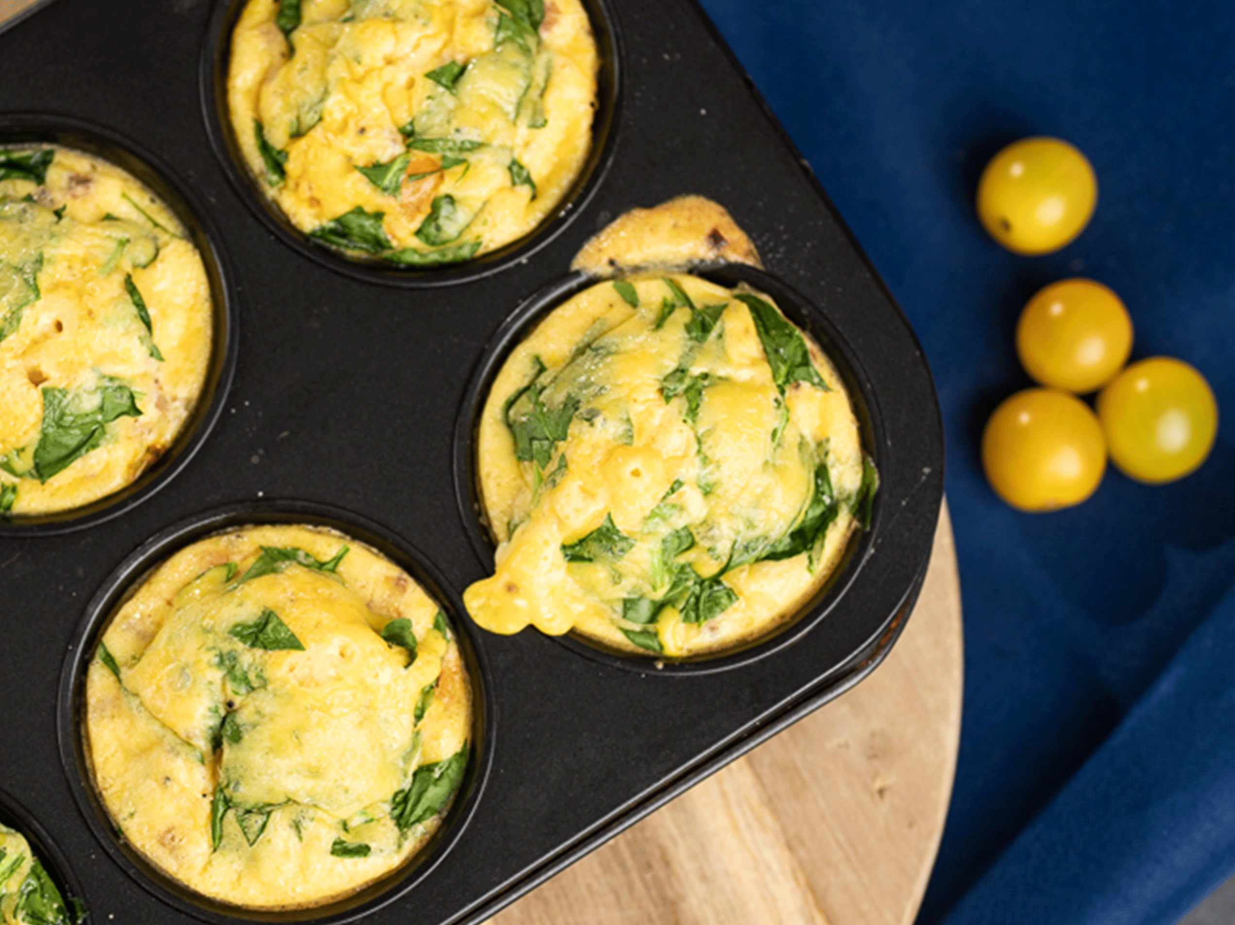 Eier-Muffins mit Spinat in einer Backform, daneben gelbe Kirschtomaten auf blauem Tuch.