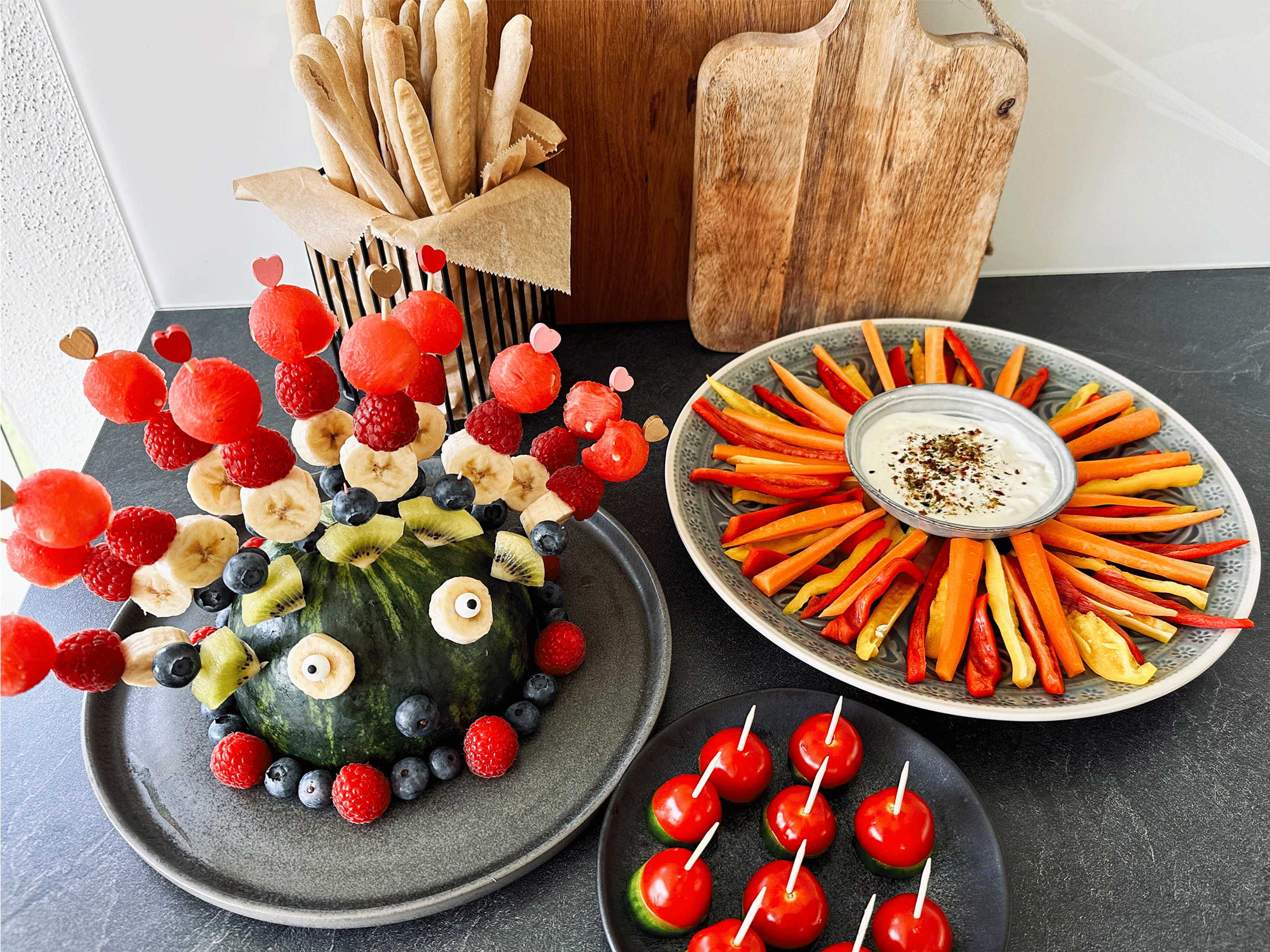 Bunter Obst-Wassermelonen-Igel mit Beeren, Bananen und Kiwis, dazu Gemüsesticks mit Dip, Grissini und Tomaten-Häppchen.
