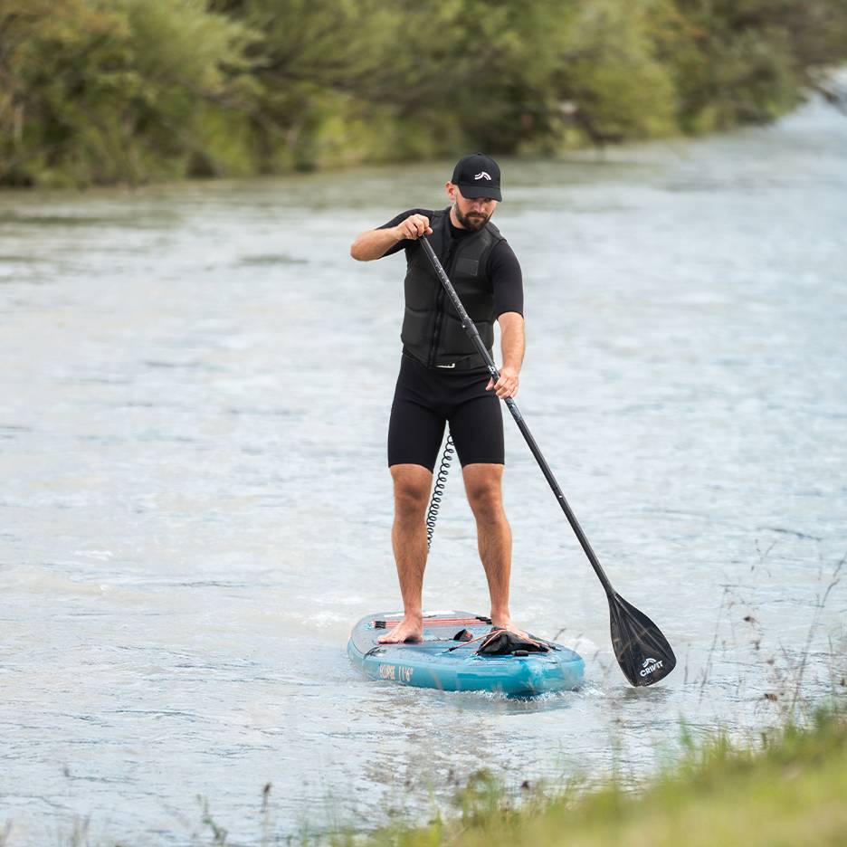 Mann beim Stand-Up-Paddling auf einem Fluss, trägt schwarzen Neoprenanzug, Schwimmweste und Kappe.