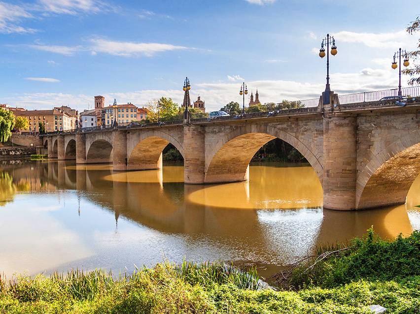 Steinbogenbrücke über einen Fluss mit einer Stadt und blauem Himmel im Hintergrund.
