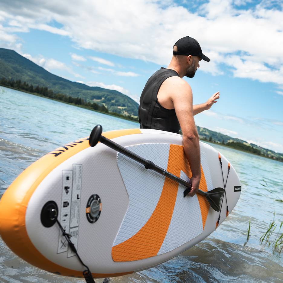 Mann mit Schwimmweste und Kappe hält aufblasbares SUP-Board im See.
