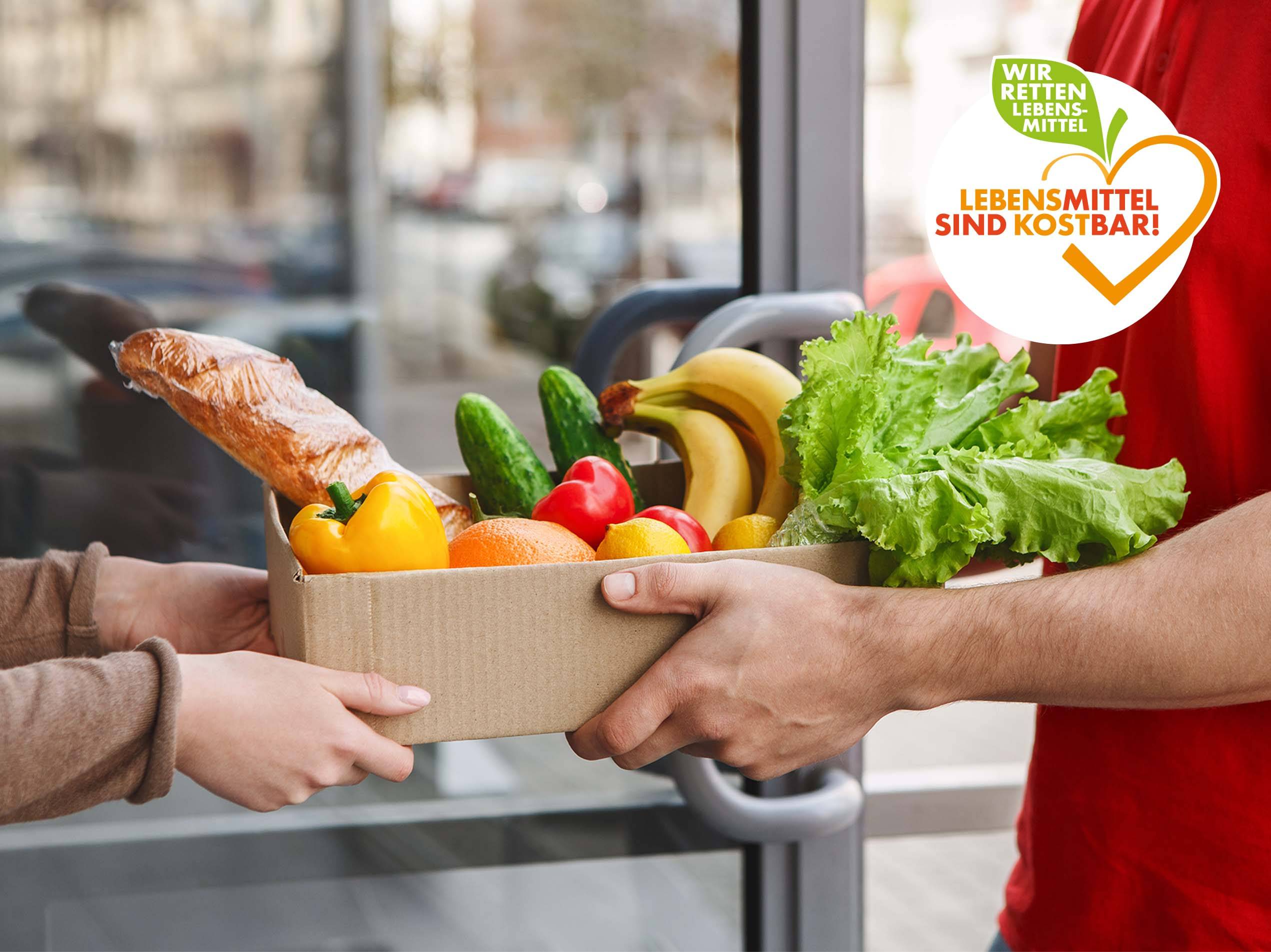 Eine Person übergibt einer anderen eine Kiste mit frischen Lebensmitteln wie Brot, Paprika, Gurken, Bananen und Salat.