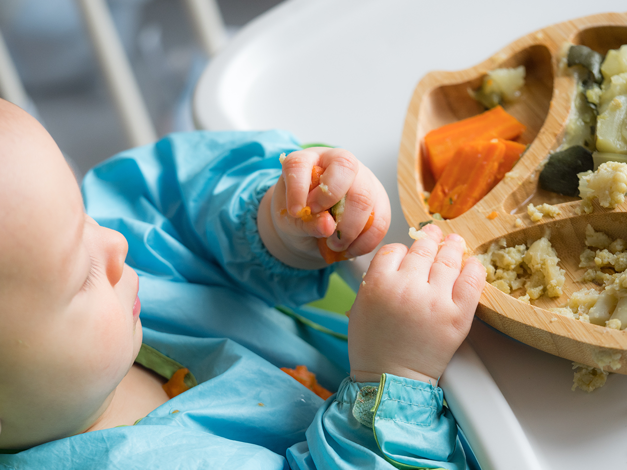 Baby in blauem Lätzchen isst Gemüse vom Holzteller