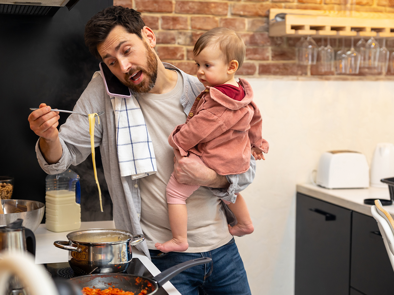 Ein Vater kocht mit Baby auf dem Arm, telefoniert und probiert Nudeln.