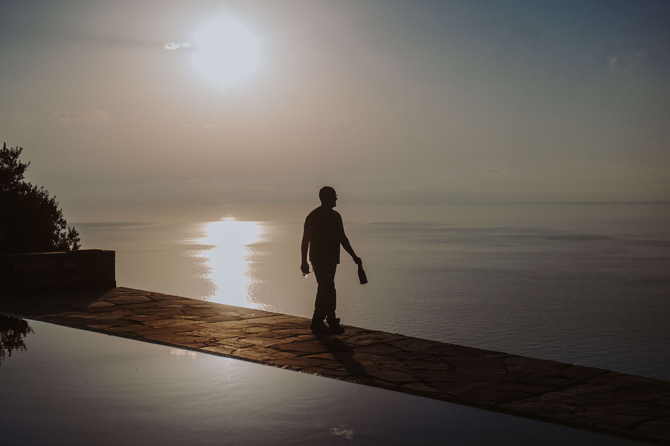 Silhouette einer Person mit Flasche und Glas an einem Infinity-Pool bei Sonnenuntergang