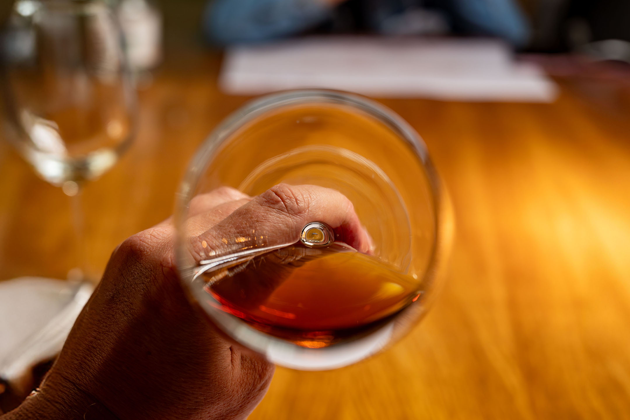 Hand hält ein Glas bernsteinfarbene Flüssigkeit, möglicherweise Brandy oder Whisky, über einem Holztisch.