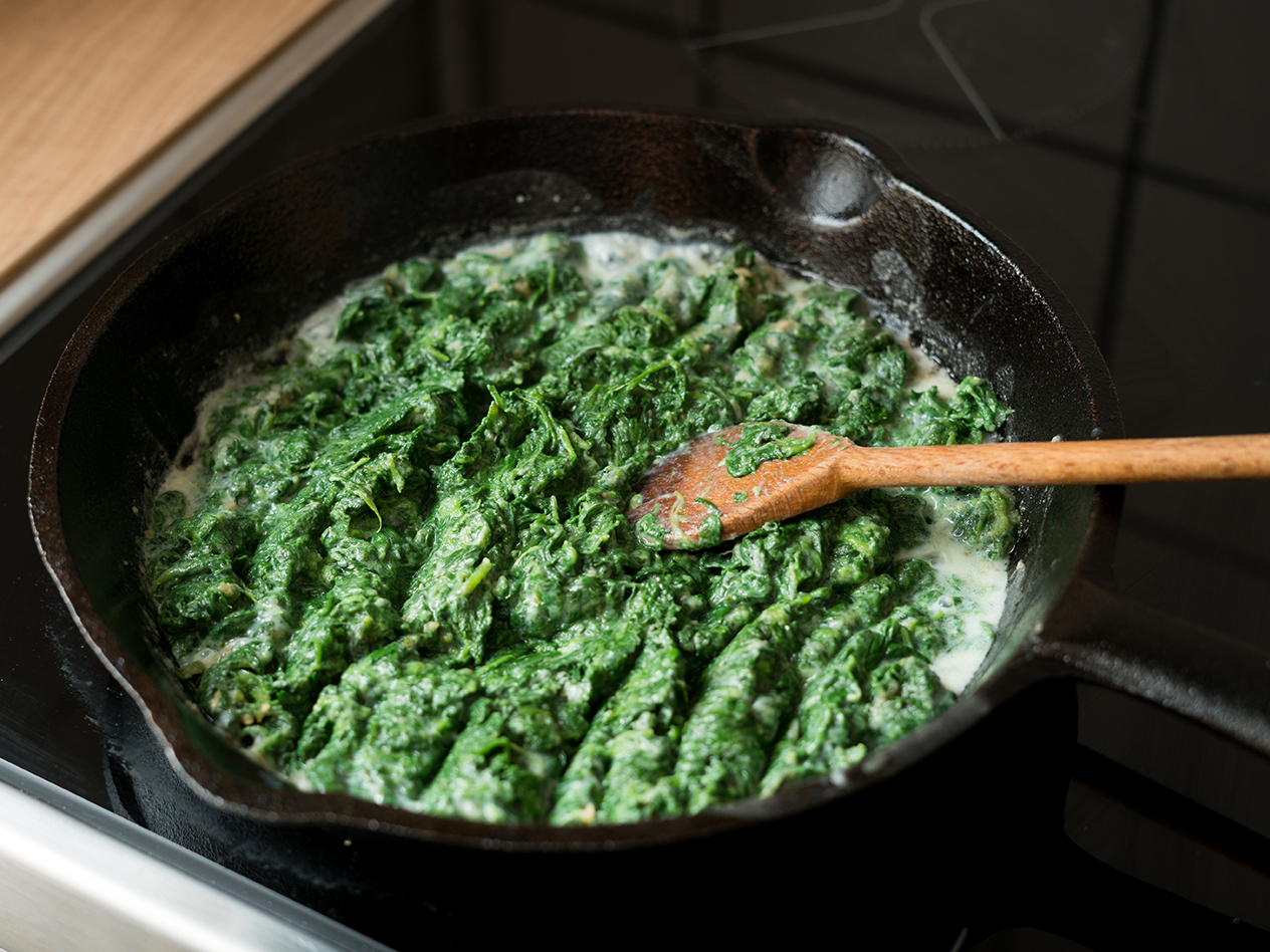 Cremiger Spinat in einer gusseisernen Pfanne mit einem Holzlöffel auf einem Herd.