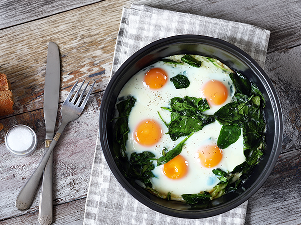 Gebackene Eier mit Spinat in einer schwarzen Pfanne auf einem Holztisch mit Besteck.