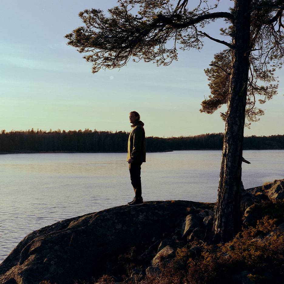 Mann in Wanderjacke an einem See, mit einem Baum im Vordergrund.