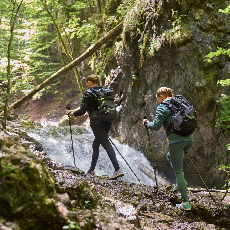 Zwei Frauen mit Rucksäcken und Trekkingstöcken wandern an einem Wasserfall entlang.