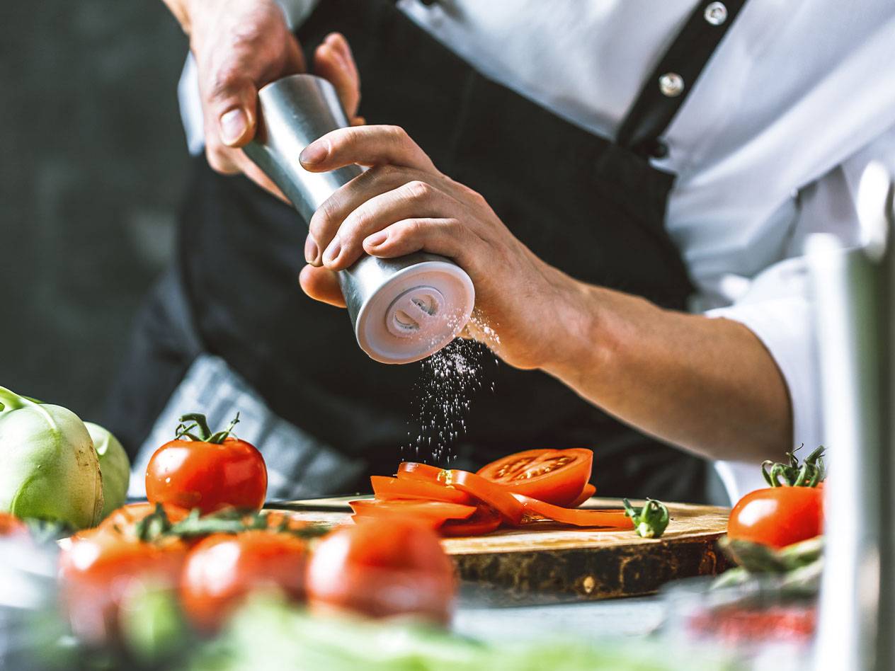 Ein Koch würzt geschnittene Tomaten mit einem Salzstreuer.
