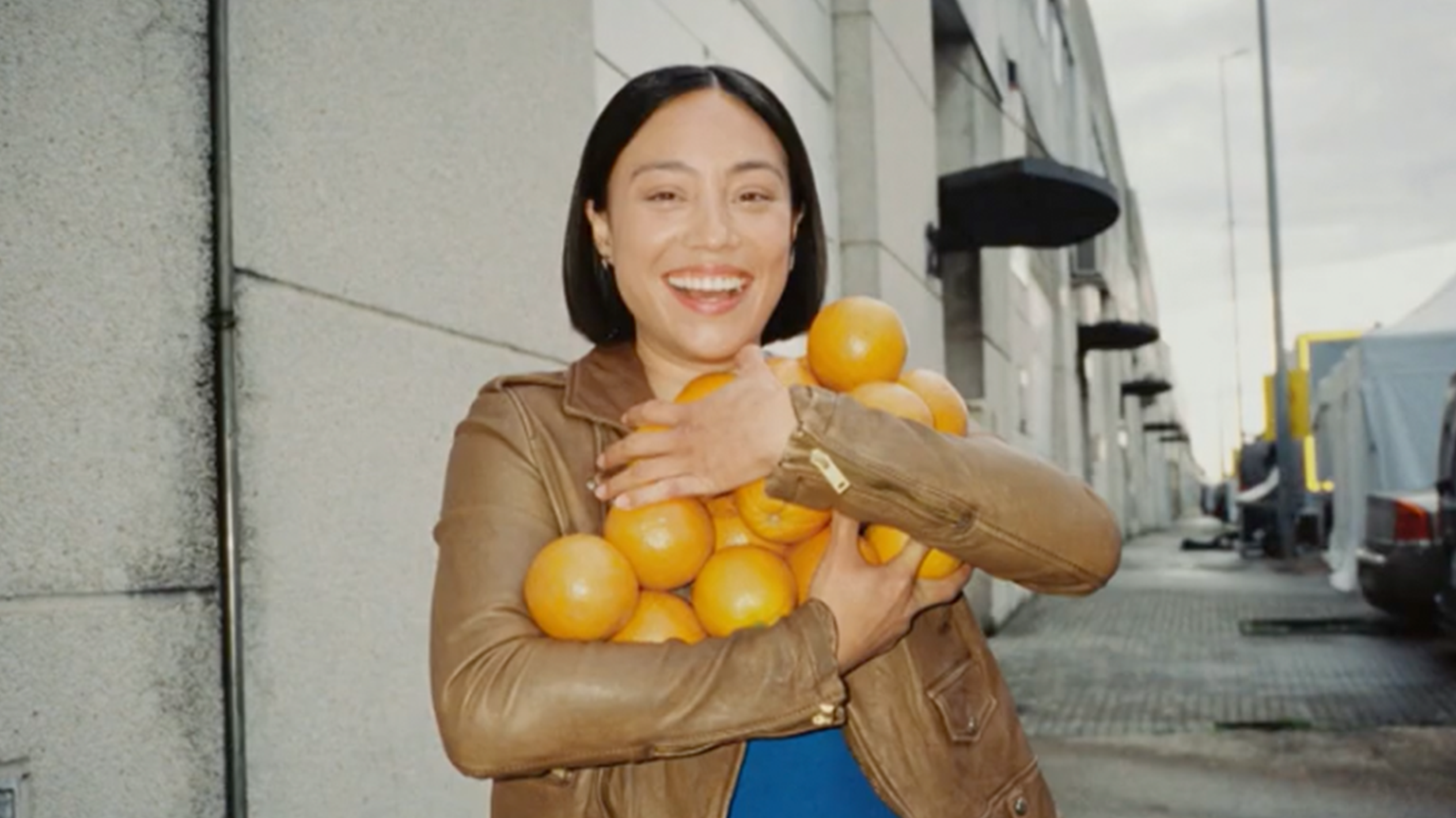 Eine lächelnde Frau in einer braunen Lederjacke hält einen Arm voller Orangen.