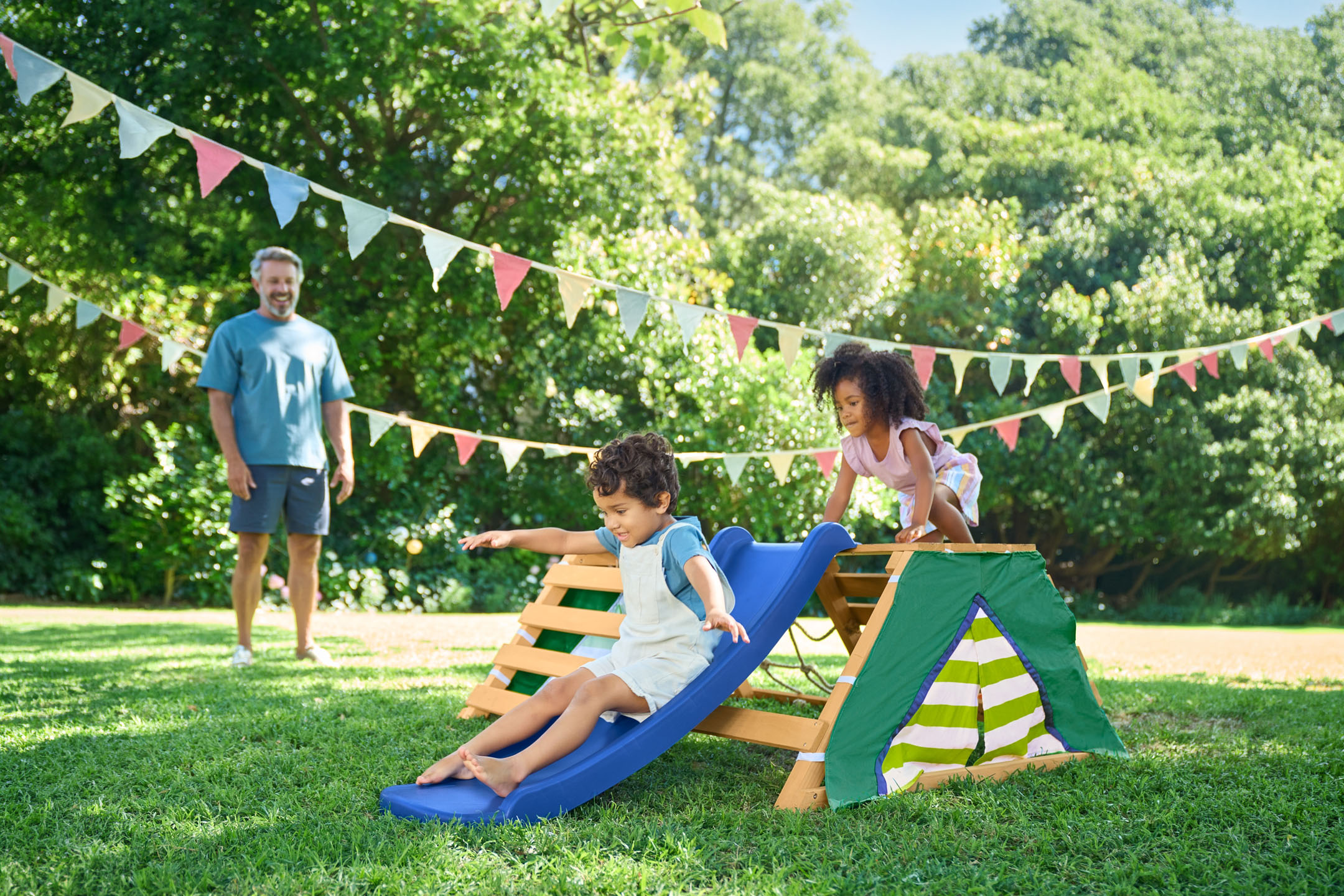Kinder spielen auf einem Holzspielplatz mit Rutsche im Garten, mit ihrem Vater im Hintergrund.