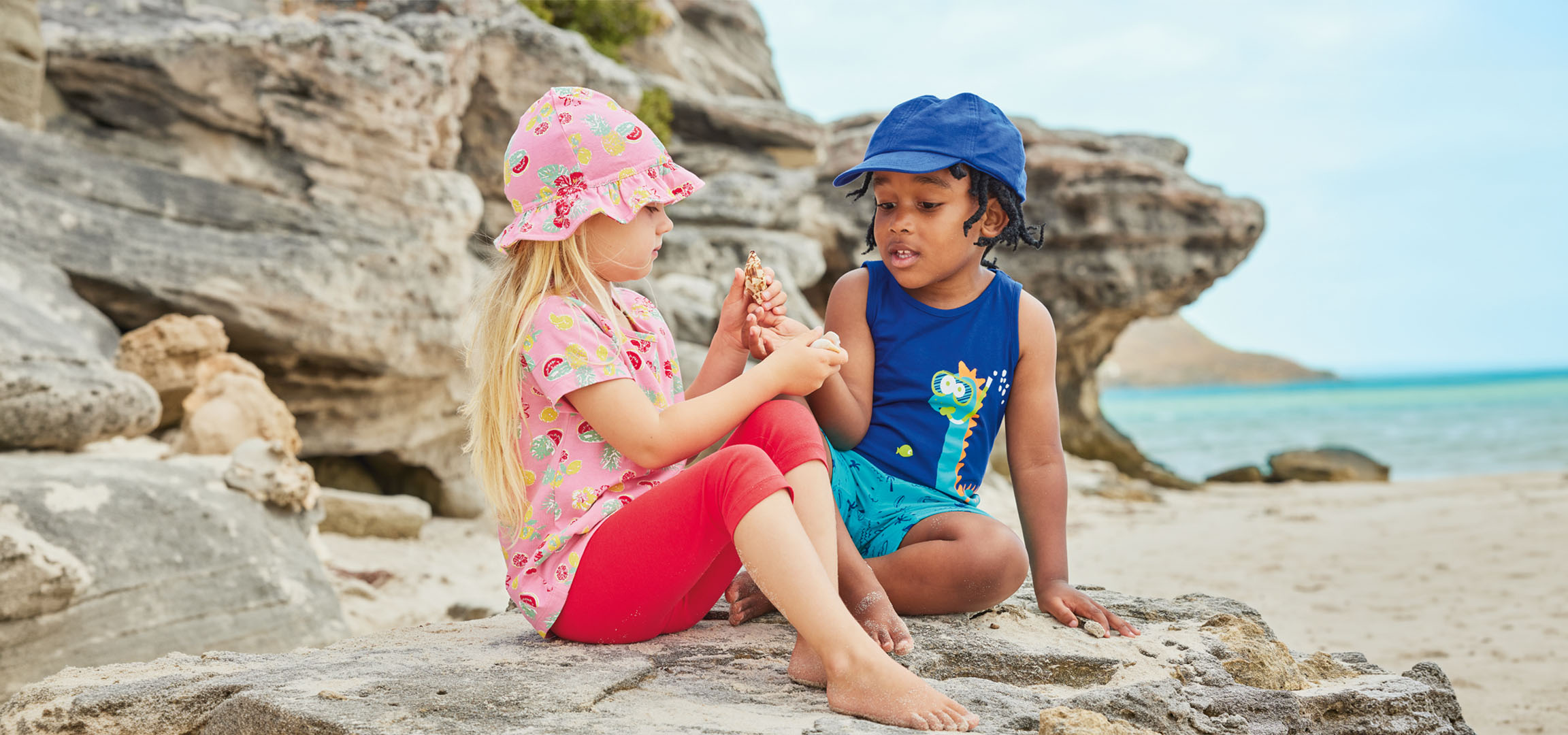 Zwei Kinder in Sommerkleidung sitzen auf einem Felsen am Strand und halten eine Muschel.