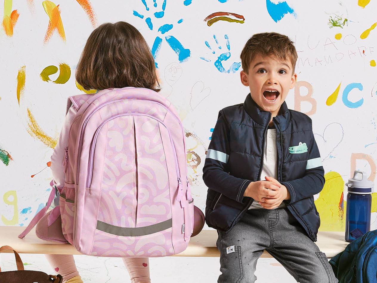 Zwei Kinder mit Schulranzen vor einer bemalten Wand; der Mädchenrucksack ist rosa mit Muster.