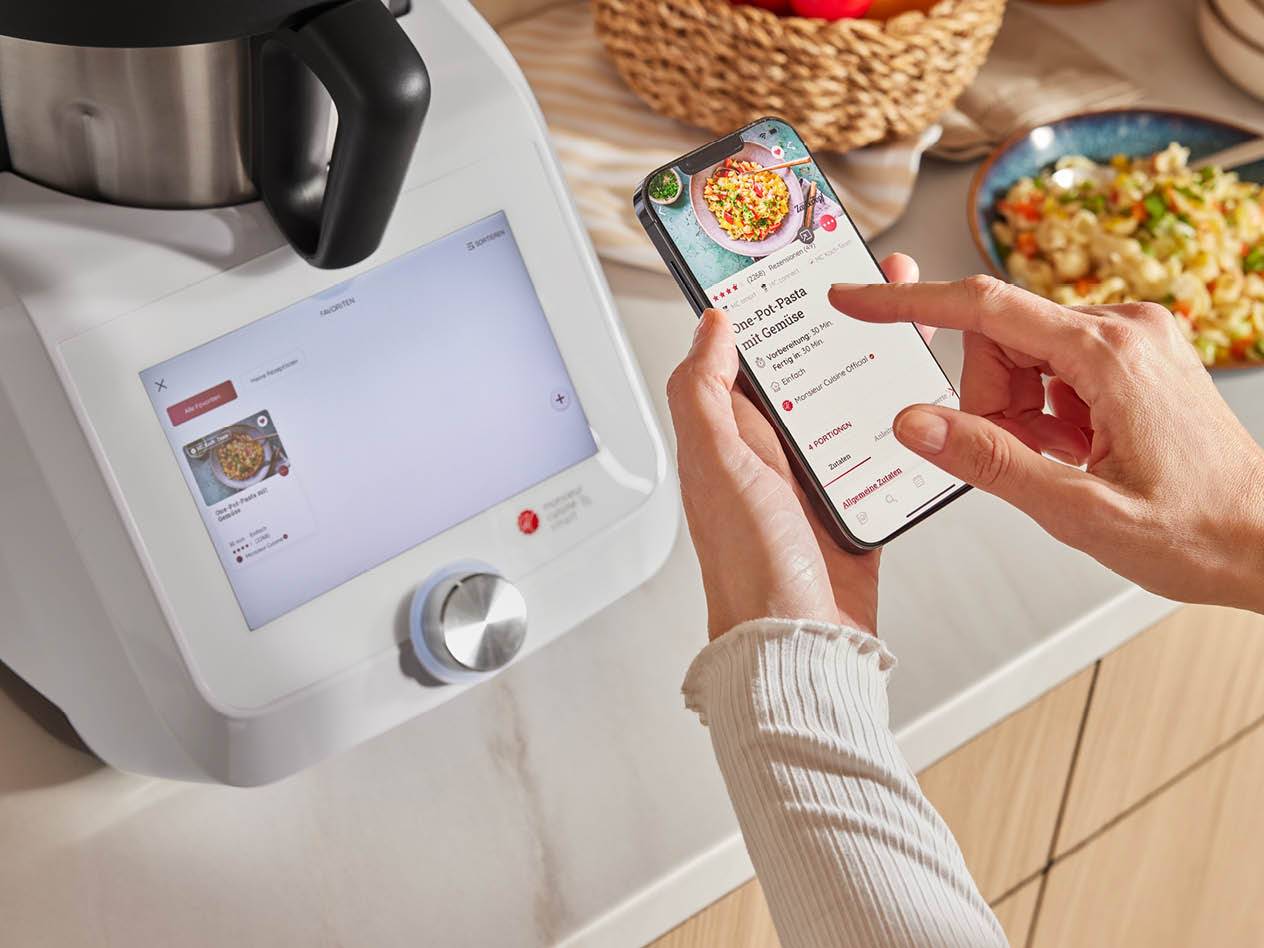 Eine Person, die ein Smartphone mit einem Rezept für One-Pot-Pasta und eine Küchenmaschine benutzt.