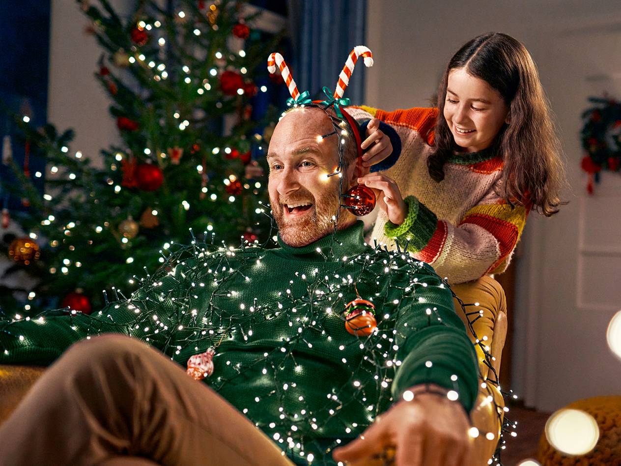 Ein Mann in Weihnachtslichtern und ein Mädchen, das ihn mit Weihnachtsschmuck schmückt.