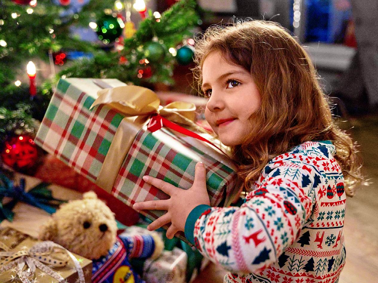 Mädchen in Weihnachtspyjama hält ein Geschenk vor einem Weihnachtsbaum mit einem Lidl-Teddybär.